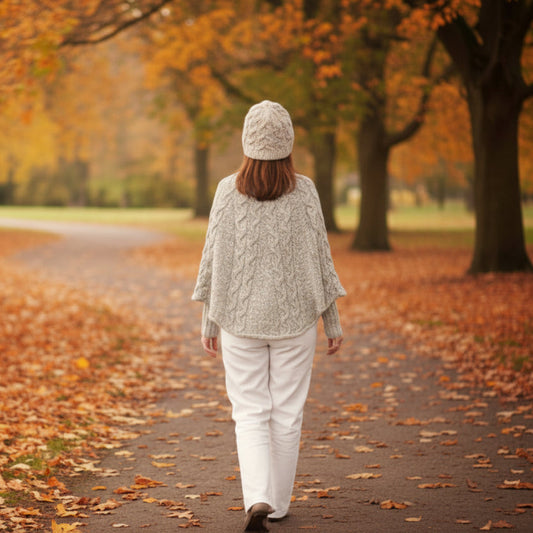 Person walking down a path wearing a beige cabled poncho with a matching hat in an autumn park with trees and fallen leaves.